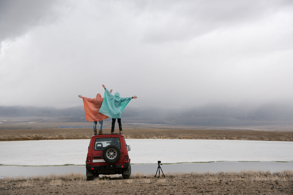 Two people in ponchos stand on top of car and look out at landscape with arms raised